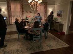 a group of people sitting around a table in a living room with curtains on the windows