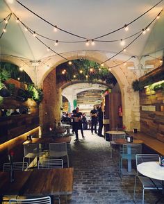 people are sitting at tables in the middle of an alleyway with lights strung over them