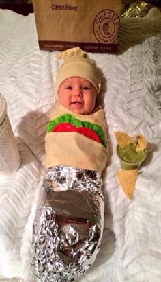 a baby is dressed up like a hot dog and wearing a taco hat while sitting on a bed