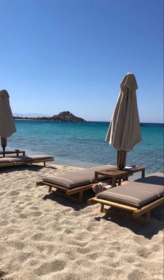 two lounge chairs and umbrellas on the beach near the blue water with an island in the background