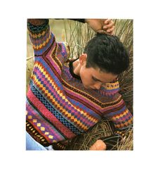 a man sitting in the grass with his hands on his hips wearing a multicolored sweater