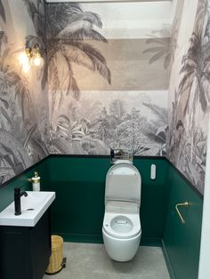 a white toilet sitting next to a sink in a bathroom under a window with palm trees on the wall