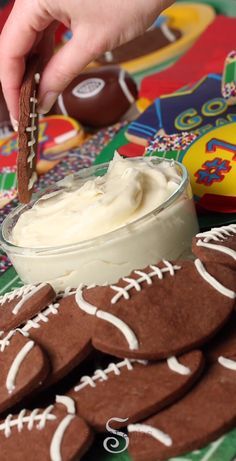 a person dipping some kind of cream in a bowl with football cookies on the table