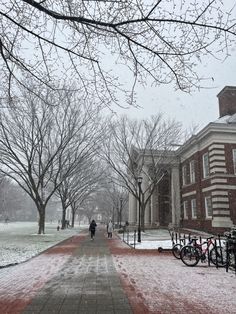 two people walking down a snow covered sidewalk in front of a brick building and trees