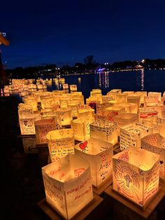 many lit up boxes are sitting on the ground near water at night with people standing around them