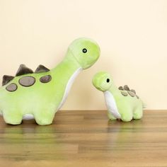 two stuffed dinosaurs sitting on top of a wooden table next to each other, one green and the other gray