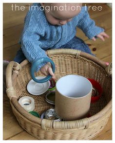 a baby sitting in a wicker basket on the floor playing with cups and saucers