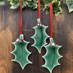 two green holly ornaments hanging from red ribbon on wooden table with greenery in background