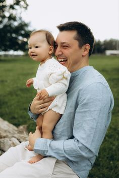 a man holding a baby in his arms while sitting on a tree stump with the grass behind him