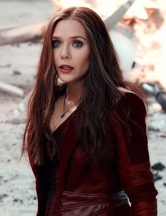 a woman with long brown hair and blue eyes wearing a red leather jacket standing in front of a fire