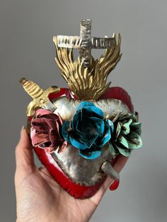 a hand holding a heart shaped object with flowers on it's side and wings