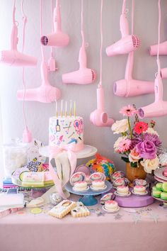 a table topped with lots of cakes and cupcakes next to pink hair dryers