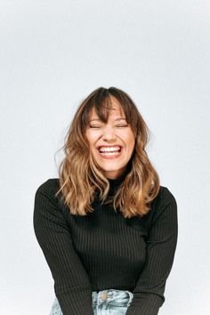 a smiling woman with her arms crossed and eyes closed, wearing a black sweater and denim skirt