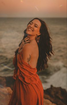 a woman in an orange dress standing on the rocks by the ocean with her arms around her neck