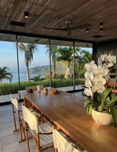 a large wooden table with white chairs and flowers on it in front of windows overlooking the ocean