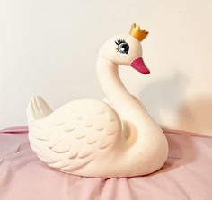 a white swan with a crown on its head sitting on a pink sheet in front of a white wall