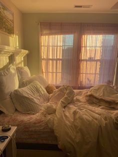 an unmade bed with white sheets and pillows in front of a window covered by curtains