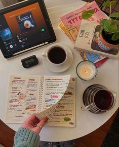 a table with a laptop, coffee cup and menus on it next to a person's hand holding an open book