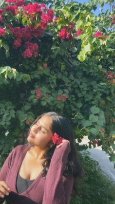 a woman is standing in front of a bush with red flowers on it and her hair blowing in the wind