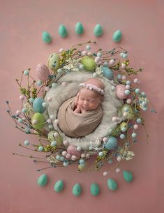 a newborn baby wrapped in a blanket surrounded by easter eggs and flowers on a pink background