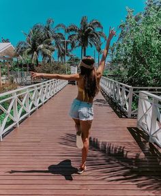 a woman walking across a wooden bridge with her arms in the air and palm trees behind her