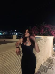 a woman in a black dress leaning against a wall at night with her hands on her hips