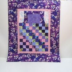 a purple and blue quilt hanging on a wall next to a white wall with flowers