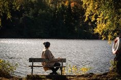 a woman sitting on a bench next to a body of water with trees in the background