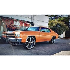 an orange muscle car parked in front of a building with black rims and chrome wheels