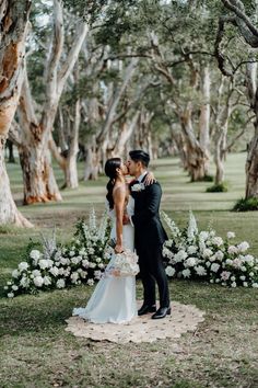 a bride and groom kissing in front of trees at their outdoor wedding ceremony with white flowers