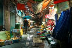 an outdoor market with lots of food and people walking through it at night time,