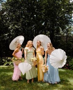 four women in dresses and umbrellas posing for the camera on grass with trees in the background