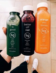 three bottles of orange, green and red juice in front of someone's feet