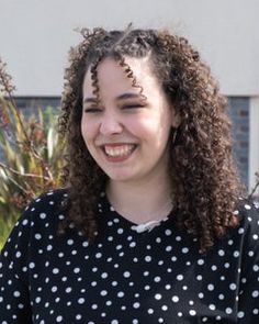 a woman with curly hair smiling and wearing a black polka dot shirt, standing in front of a palm tree