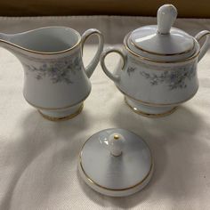 three pieces of white porcelain with gold trimmings and flowers on the sides, including a creamer and sugar bowl