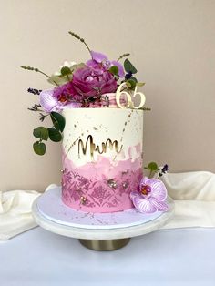 a pink and white cake with purple flowers on the top is decorated with gold letters