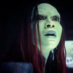 a woman with long red hair and green makeup in a dark room, looking at the camera