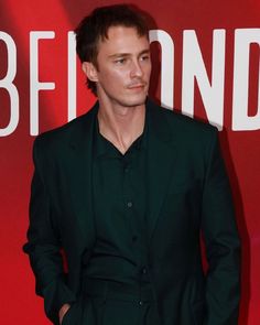 a man in a black suit standing next to a red wall with the words behind him