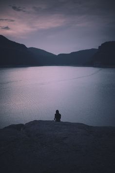 a person sitting on top of a rock next to a body of water under a cloudy sky