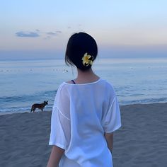 a woman standing on top of a sandy beach next to the ocean with a yellow flower in her hair