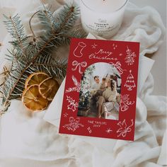 a christmas card sitting on top of a white blanket next to a candle and some pine branches