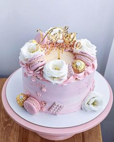 a pink birthday cake with white flowers and gold letters on top is sitting on a table