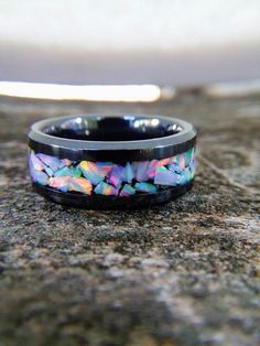 a black ring with multicolored opal inlays sitting on top of a rock