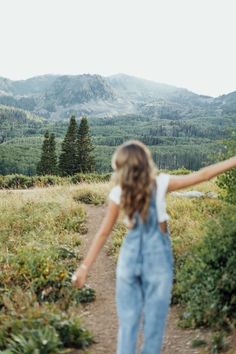 a girl in overalls walking down a dirt path with her arms outstretched towards the mountains