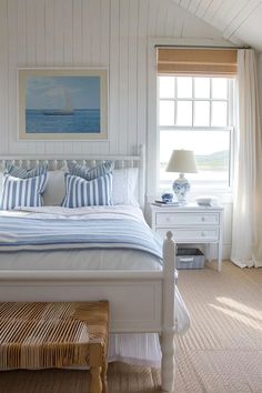 a white bed with blue and white pillows in a bedroom next to a painting on the wall