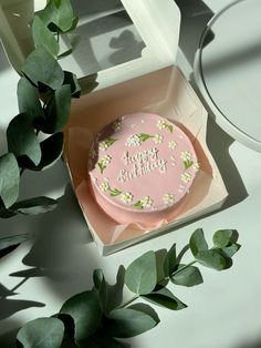 a pink birthday cake in a box with flowers on the side and greenery around it