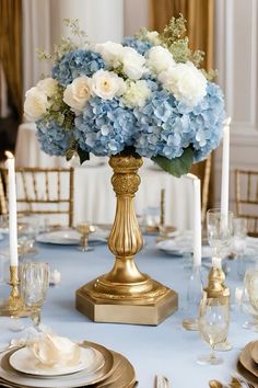 blue and white flowers in a gold vase on top of a table with silverware