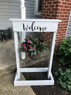 a welcome sign sitting in front of a house with potted plants on the side