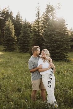 a man and woman hugging in the middle of a field with pine trees behind them