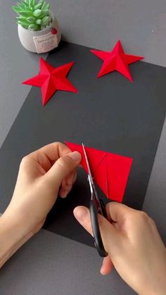 someone cutting out red paper stars on top of a gray surface with scissors and a potted plant in the background